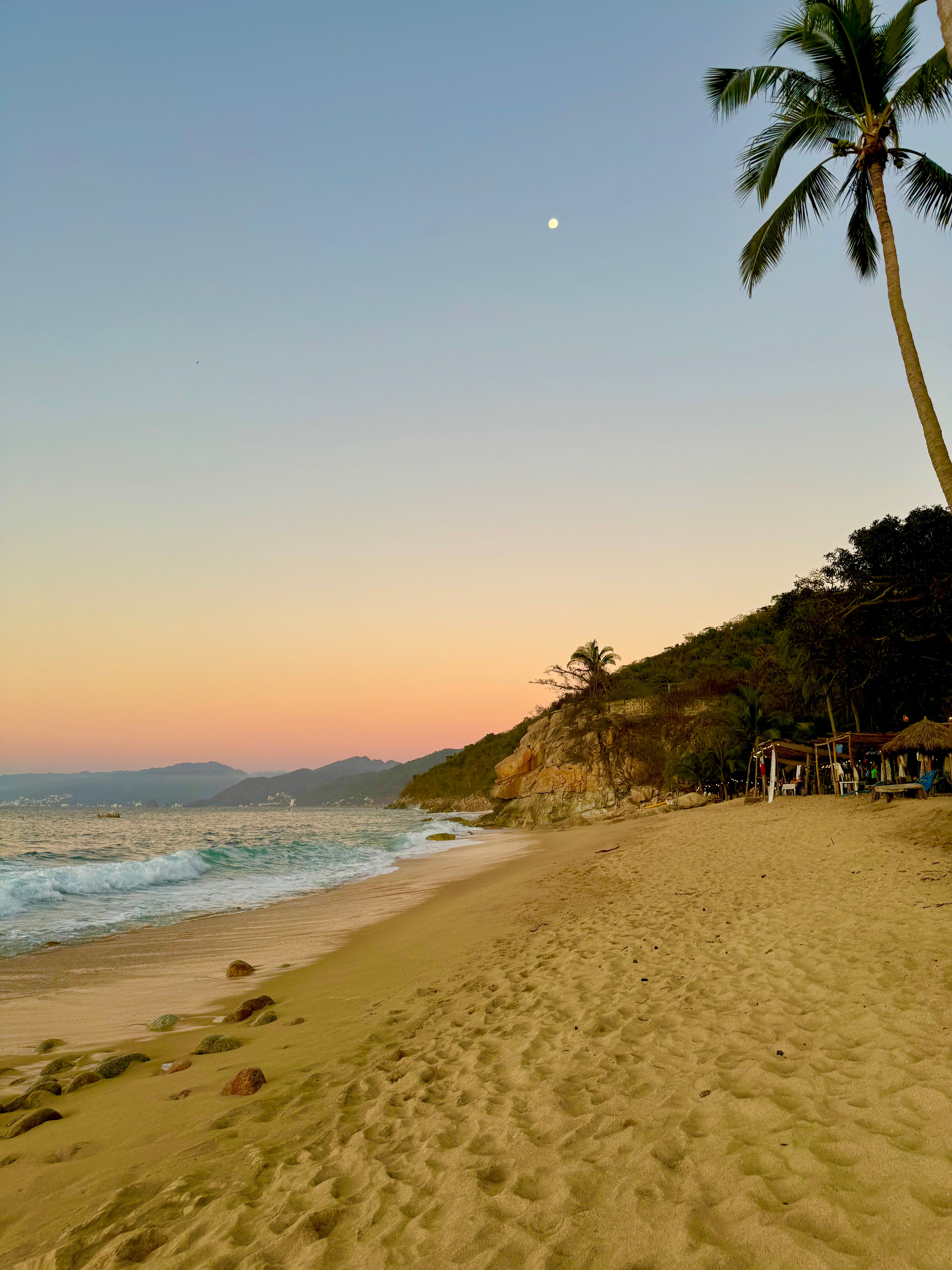 Beach at dusk with surf, palm trees, and cliffside thatched cabanas